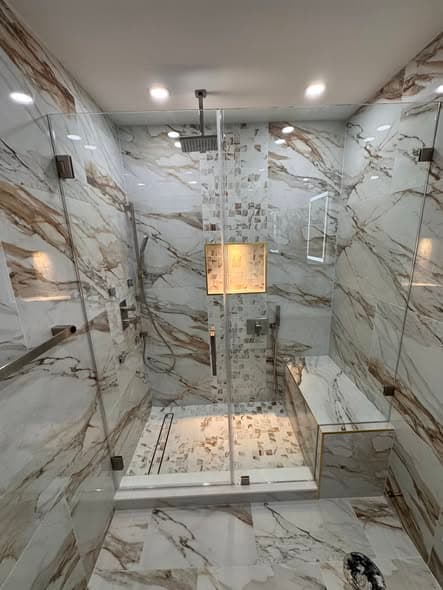 Frameless glass shower enclosure with calacatta marble-look porcelain tile walls featuring brown and gold veining, mosaic tile shower floor and accent niche, ceiling-mounted rain showerhead, and built-in bench in a Bridgewater NJ bathroom remodel